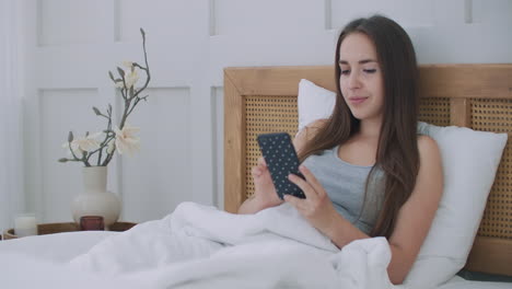 Woman-holding-mobile-phone-while-lying-in-bed-at-home.